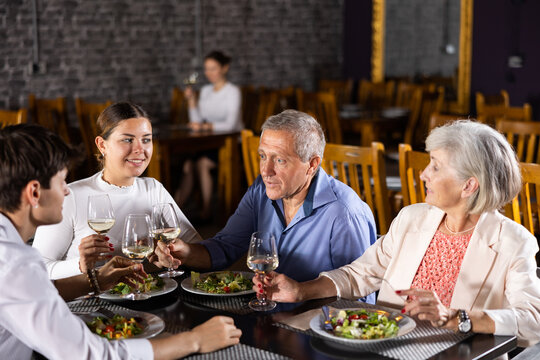 Happy Senior Woman And Man Meeting With Adult Children Over Dinner In Restaurant. People Talking Cheerfully And Drinking Wine At Table