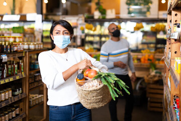 Fototapeta Female shopper wearing protective mask with bag of groceries and vegetables at grocery supermarket obraz