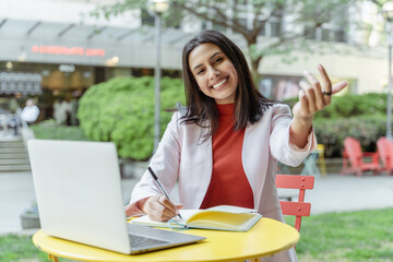 Smiling smart businesswoman studying, learning language, listening webinar sitting outdoors. Portrait of successful asian woman, young writer taking notes in cafe 