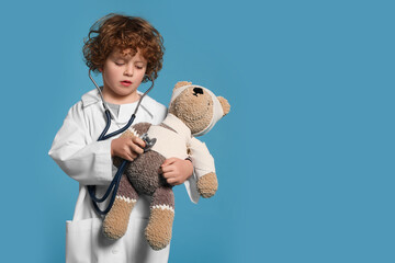 Little boy in medical uniform examining toy bear with stethoscope on light blue background. Space...