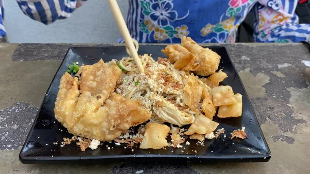 Hands stirring fried noodles with pangsit wonton topping using chopsticks