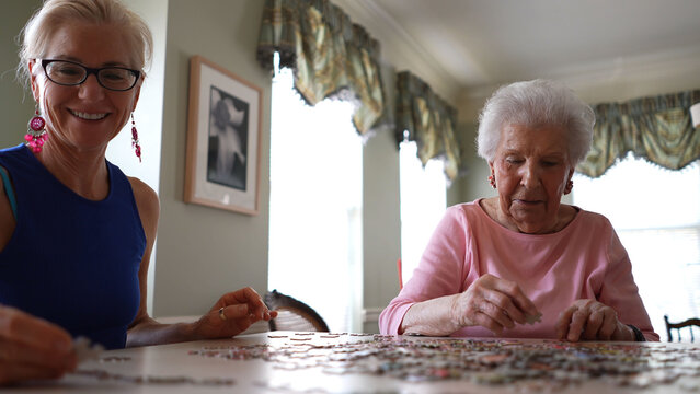 Smiling mature woman places puzzle piece while elderly lady moves puzzle pieces around. Concept of spending time with loved ones.