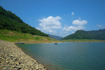 Upstream view on the dam's crest with a long-tailed boat. Surrounded by mountains and lush nature.