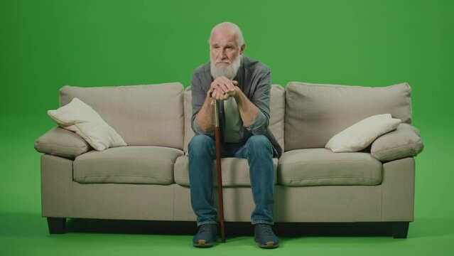 Green Screen. A Sad Old Man with a Stick Is Sitting On A Sofa. An Elderly Man Leaning His Head On A Stick and Rubs His Gray Beard. Mental Health for Seniors.