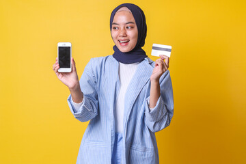 Excited young Asian Muslim girl showing blank screen of mobile phone for copy space while holding credit card isolated over yellow background. Online payment 
 business concept. 