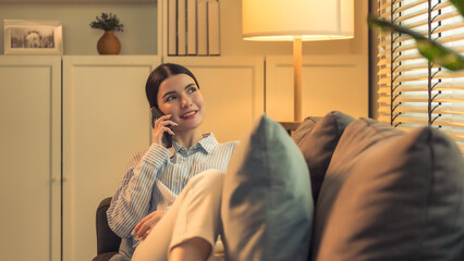 Fototapeta premium this portrait captures a beautiful young asian woman, engaging in a phone call while leisurely leaning back on a couch in a cozy, warmly lit living room