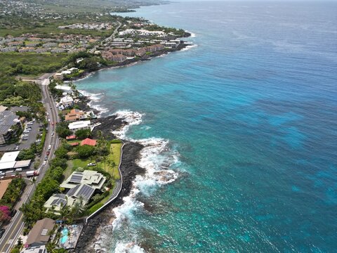 View of coastal Kailua-Kona on the Big Island of Hawai'i. Aerial drone looking along the coast with palm trees, Condominiums, coastal highway, and blue and green waters with volcanic rock and coral.