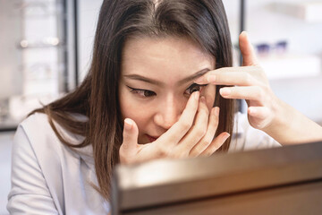 Asian young woman putting contact lenses into her eyes at optical shop, trying on new contact lenses, selective focus