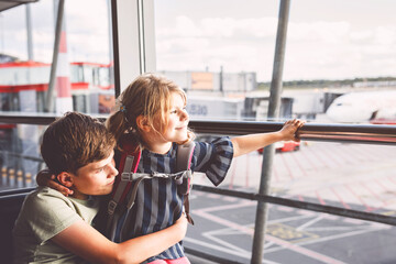 Little girl and school boy at the airport waiting for boarding at big window. Two kids stands at window against the backdrop of airplanes. Happy children, siblings leaving for family summer vacation