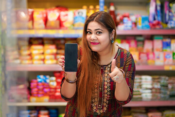 Happy indian woman showing smartphone screen with thumps up at grocery shop.