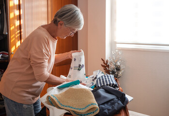 Smiling mature lady packing her stuff into a suitcase preparing for vacation trip. Elderly woman checking her luggage on the bed at home before to leave