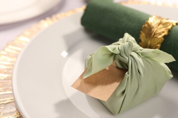 Furoshiki technique. Gift packed in green fabric, blank card and napkin on plate, closeup. Space for text