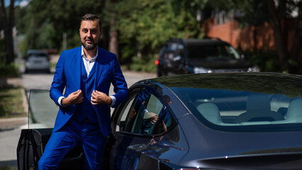Fototapeta na wymiar Caucasian bearded man in a blue suit gets out of a black electro car in the countryside in summer.