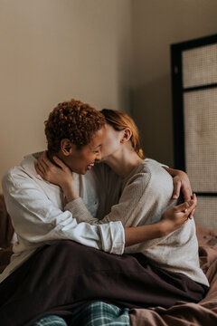 Non-binary person kissing friend while sitting on bed at home