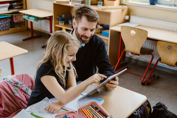Smiling female student using tablet PC held by male teacher in classroom