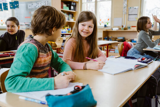 Smiling girl talking to male friend while sitting at desk in classroom