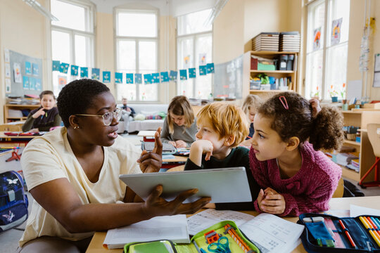 Teacher with tablet PC explaining schoolboy and schoolgirl at desk while gesturing in classroom