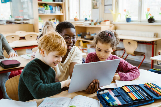Happy teacher assisting boy and girl with digital tablet sitting at desk in classroom