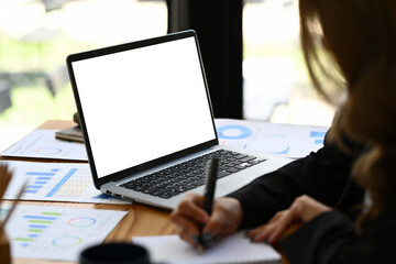 Cropped shot of female employee sitting at office desk with laptop and document, writing on notebook