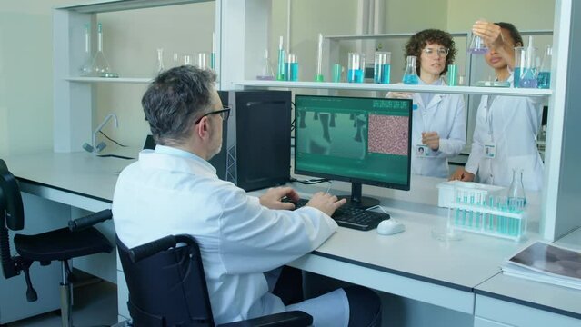 Arc shot of male professor in wheelchair approaching desk to work with computer while two Caucasian and Biracial lab assistants having experiment with flasks