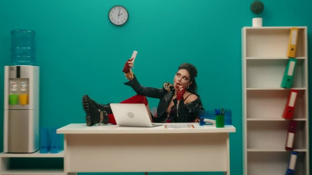 A woman freak, being at the workplace, takes a selfie on a smartphone and eats a lollipop. A woman with bright makeup sits in the office with her feet on the table.Advertising.