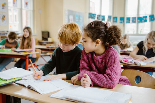 Girl sitting besides blond boy writing in book at desk in classroom - Powered by Adobe