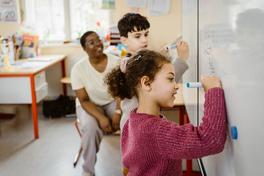 Girl and boy writing on whiteboard while solving mathematics in classroom - Powered by Adobe