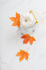 white halloween pumpkin in little grocery trolley with skeleton arms and maple leaves on marble background