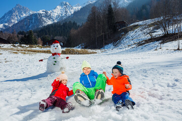 Three children jump together in snow near snowman over mountains