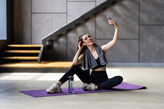 Young fitness trainer instructor woman doing exercises do selfie shot on mobile cell phone sit on mat floor at home