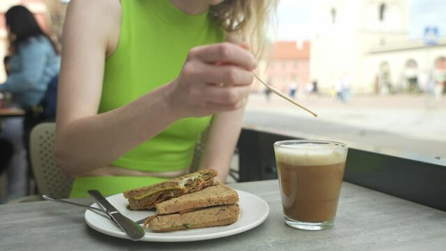 Woman sits in the coffee shop terrace and drinks cold cappuccino with caramel. Crispy sandwich on the table. Summer lunch in the city. Female mixing her cold coffee before drinking it.