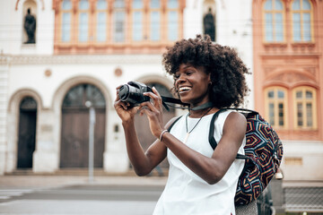 Young black female tourist enjoys walking through the streets of a beautiful European city. She is happy and using her photo camera to take fantastic architecture photographs. Bright sunny day. - obrazy, fototapety, plakaty