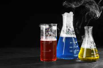 Laboratory glassware with colorful liquids and steam on dark table against black background, space for text. Chemical reaction
