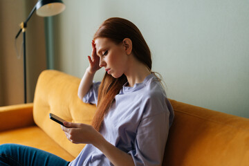 Close-up side view of sad young woman sitting on yellow couch looking at mobile phone screen,...