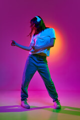 Portrait of young girl wearing casual clothes and headphones listening to music with joy and moving in beat of song over pink background in neon light