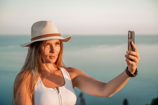 Selfie woman in hat, white tank top and shorts makes selfie shot mobile phone post photo social network outdoors on sea background beach people vacation lifestyle travel concept.