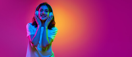 Positive brunette woman wearing casual clothes and headphones touching cheeks, smiling over pink studio background in neon light. DJ girl