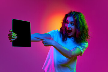 Beautiful girl, student in stylish clothes holding tablet and pointing at empty screen over pink background in neon light. Concept of online school, emotions