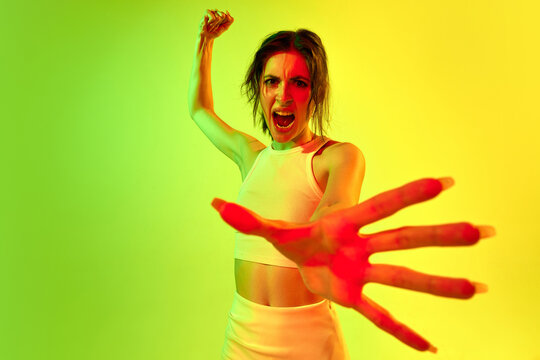 One angry woman swinging fists and screaming with open mouth over acid green color studio background in neon light. Concept of challenges, human emotions