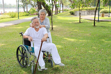 Asian senior woman or caregiver helping and consoling senior man walk with wheelchair at park outdoor. Elderly wife taking good help care and support of elder husband patient outside the house.