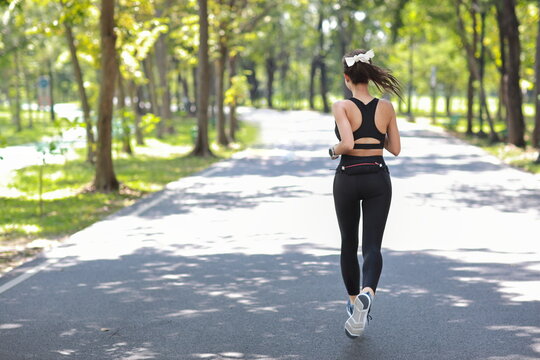 Rear view asian fitness female sportswoman runner doing morning jogging exercise training outdoor. Happy smile athlete woman running under the tree in the park while looking at camera and listen music