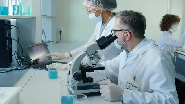 Dolly zoom of middle aged male Caucasian scientist sitting in wheelchair at desk looking in microscope talking to Biracial female colleague looking at screen with cells