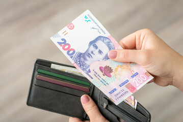 Fototapeta Banknote of 200 hryvnias in female hand close up. Grey wallet with ukrainian money and credit cards on the background, paying in cash obraz