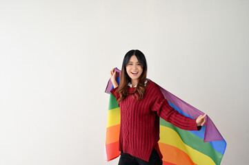 A pretty Asian woman with a rainbow LGBT flag stands over an isolated white studio background.
