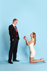Young beautiful woman in white dress standing on knees, proposing to her boyfriend to get married against blue studio background. Concept of family, relationship, youth, emotions, fun. Ad