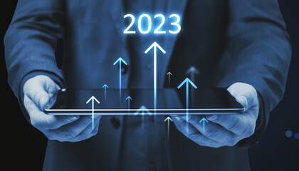 Toned businessman hands holding tablet with growing upward 2023 arrows on dark background. Company growth, annual report and increase concept.