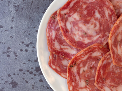 Top close view of several slices of sweet sopressata on a white plate atop a gray mottled background.