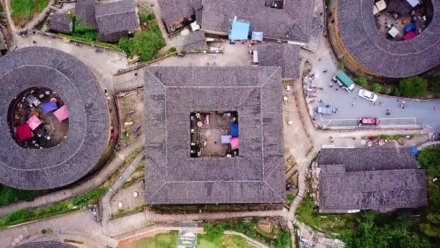 Drone shot zooms out of a square-shaped Unesco World Heritage Tulou Hakka. Traditional architecture with other Tulou houses on a mountain hill with people walking around, Fujian, China