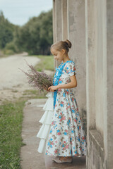 Young cute girl in vintage elegant old-fashioned dress standing sideways with bouquet of heather flowers near wall of old mansion in countryside, summer child portrait in retro village provence style