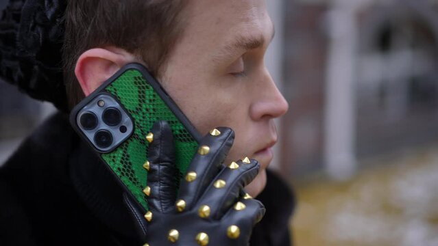 Headshot of concentrated intelligent young Caucasian man listening talking on phone standing on urban city street. Close-up portrait of gay person in stylish leather gloves with live camera panning